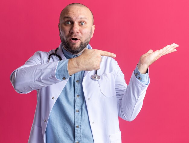 Impressed middle-aged male doctor wearing medical robe and stethoscope  showing empty hand pointing at it isolated on pink wall