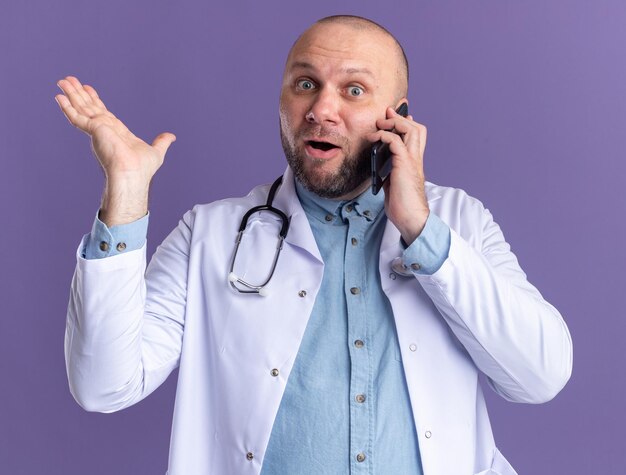 보라색 벽에 고립 된 빈 손을 보여주는 전화 통화에 의료 가운과 청진기를 입고 감동 중년 남성 의사