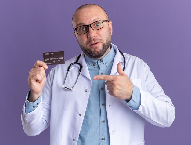 Impressed middle-aged male doctor wearing medical robe and stethoscope with glasses holding and pointing at credit card looking at front isolated on purple wall