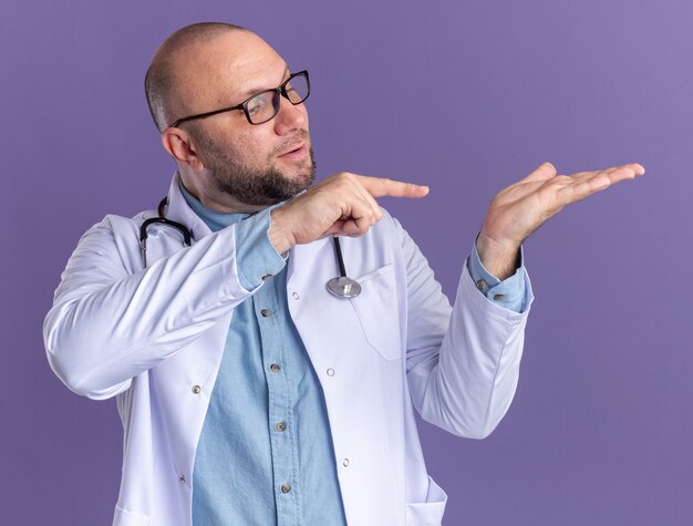 Impressed middle-aged male doctor wearing medical robe and stethoscope with glasses showing empty hand looking and pointing at it isolated on purple wall