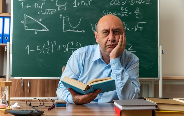 impressed middle-aged male teacher sits at table with school supplies holding book putting hand on cheek in classroom