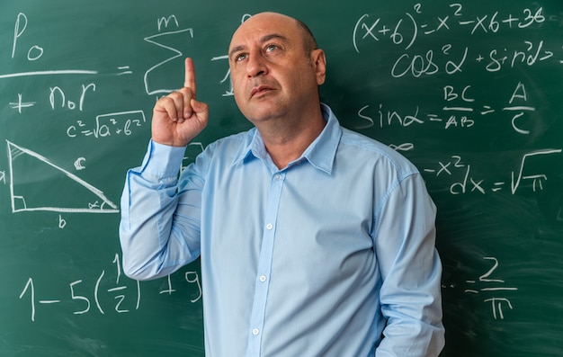 impressed middle-aged male teacher standing in front blackboard points at up