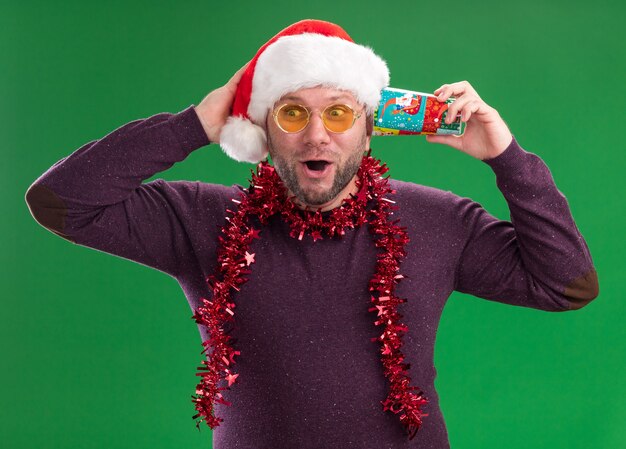 Impressed middle-aged man wearing santa hat and tinsel garland around neck with glasses holding plastic christmas cup next to ear listening to secrets keeping hand