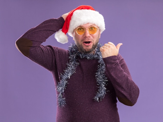 Impressed middle-aged man wearing santa hat and tinsel garland around neck with glasses  keeping hand on head pointing at side isolated on purple wall