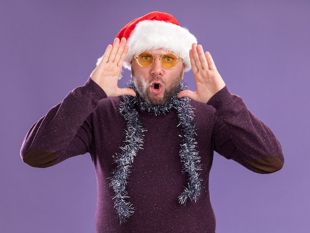 Impressed middle-aged man wearing santa hat and tinsel garland around neck with glasses  keeping hands near head isolated on purple wall