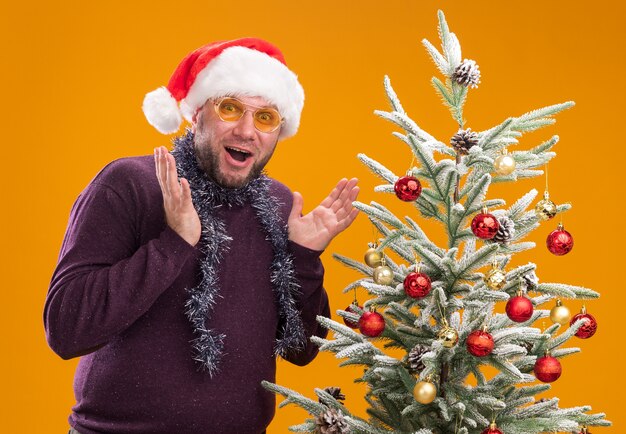 Impressed middle-aged man wearing santa hat and tinsel garland around neck with glasses standing near decorated christmas tree