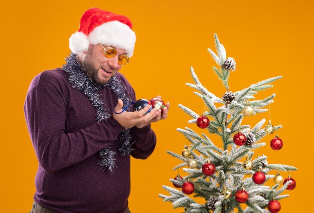 Impressed middle-aged man wearing santa hat and tinsel garland around neck with glasses standing in profile view near christmas tree holding and 