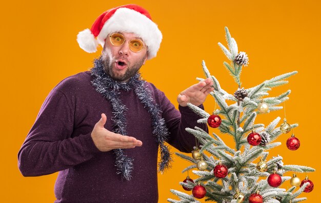 Impressed middle-aged man wearing santa hat and tinsel garland around neck with glasses standing in profile view near decorated christmas tree pointing at it 
