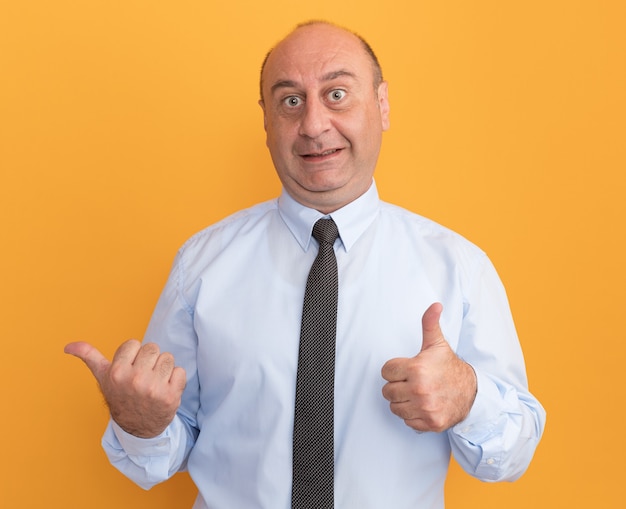 Impressed middle-aged man wearing white t-shirt with tie showing thumb up and points at side isolated on orange wall