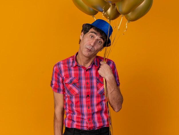 Impressed middle-aged party man wearing party hat holding balloons looking at front isolated on orange wall