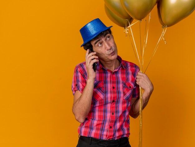 オレンジ色の壁に隔離された側を見て電話で話している風船を持ってパーティーハットを身に着けている印象的な中年のパーティー男