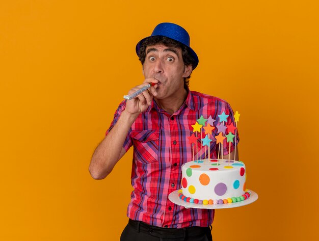 Foto gratuita impressionato uomo di mezza età del partito che indossa il cappello del partito che tiene la torta di compleanno che soffia il ventilatore del partito che esamina la parte anteriore isolata sulla parete arancione
