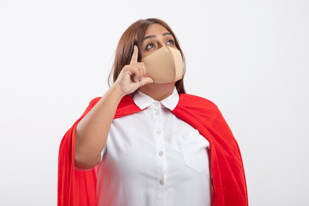 Impressed middle-aged superhero female looking up wearing medical mask putting hand on cheek isolated on white