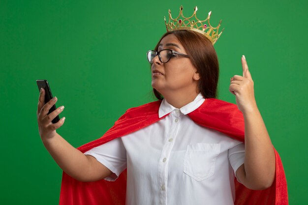 Impressed middle-aged superhero female wearing crown with glasses holding and looking at phone points at up isolated on green background