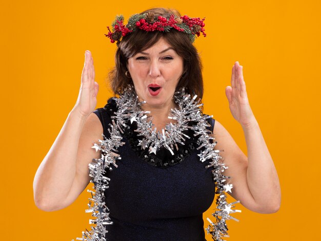 오렌지 벽에 고립 된 크기 제스처를하고 목 주위에 크리스마스 머리 화환과 반짝이 화환을 입고 감동 중년 여성