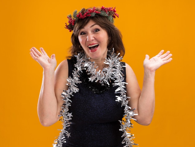 Impressed middle-aged woman wearing christmas head wreath and tinsel garland around neck showing empty hands isolated on orange wall