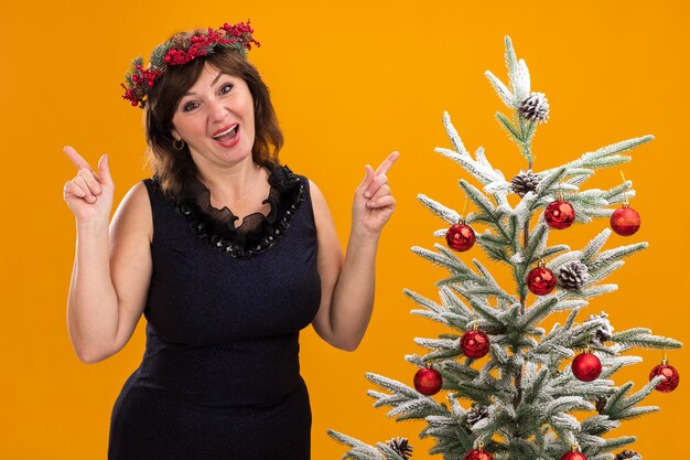 Impressed middle-aged woman wearing christmas head wreath and tinsel garland around neck standing near decorated christmas tree