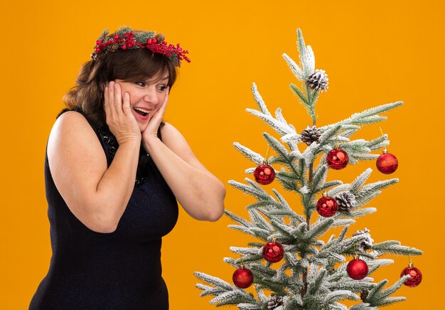 Impressed middle-aged woman wearing christmas head wreath and tinsel garland around neck standing in profile view near decorated christmas tree 