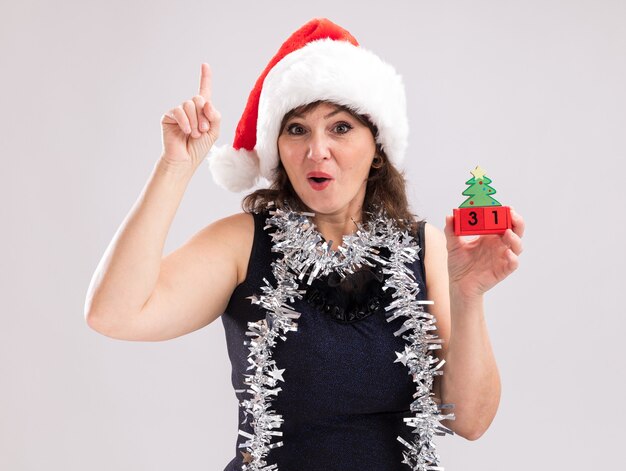 Impressed middle-aged woman wearing santa hat and tinsel garland around neck holding christmas tree toy with date looking at camera pointing up isolated on white background