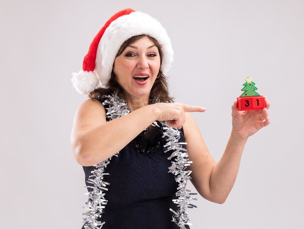 Free photo impressed middle-aged woman wearing santa hat and tinsel garland around neck holding and pointing at christmas tree toy with date looking at camera isolated on white background