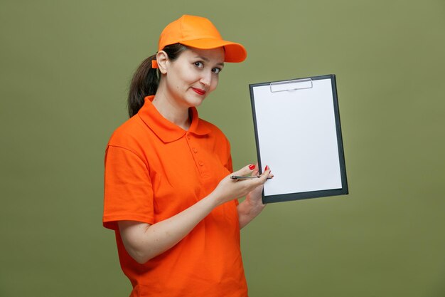 Впечатленная женщина-доставщик средних лет в униформе и кепке с ручкой, смотрящая в камеру, показывающая буфер обмена, изолированный на оливково-зеленом фоне