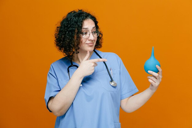 Foto gratuita impressionato dottoressa di mezza età che indossa occhiali uniformi e uno stetoscopio intorno al collo tenendo il clistere guardando e indicandolo isolato su sfondo arancione
