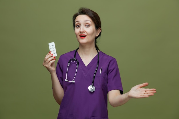 Impressed middleaged female doctor wearing uniform and stethoscope around neck showing pack of tablets looking at camera showing empty hand isolated on olive green background