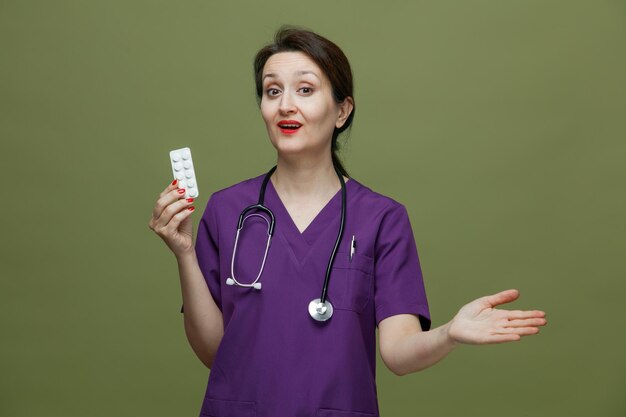 Впечатленная женщина-врач средних лет в униформе и со стетоскопом на шее, показывающая пачку таблеток, смотрящая в камеру, показывающая пустую руку, изолированную на оливково-зеленом фоне