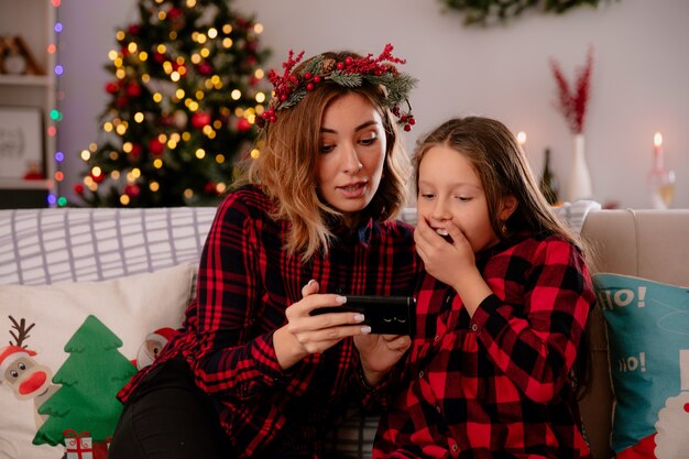 ソファに座って、家でクリスマスの時間を楽しんで電話で何かを見て感動した母と娘