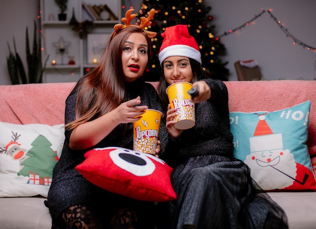 Free photo impressed mother holds popcorn bucket pointing at camera and pleased daughter with santa hat holds tv remote sitting on couch enjoying christmas time at home