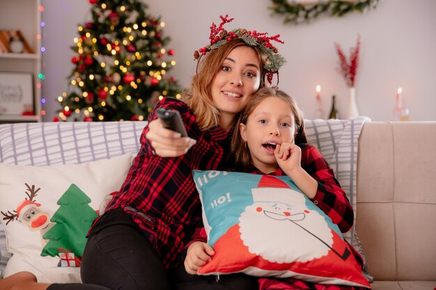 Impressed mother with holly wreath holds tv remote and looks at camera with daughter sitting on couch and enjoying christmas time at home