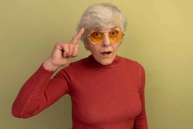 Бесплатное фото Впечатленная старуха в красном свитере с высоким воротом и солнцезащитных очках, делающая мысленный жест изолирована на оливково-зеленой стене с копией пространства