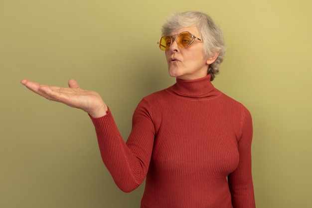 Impressed old woman wearing red turtleneck sweater and sunglasses showing empty hand looking at it 