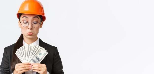 Impressed and pleased female asian construction inspector industrial architect receiving big bribe counting money and looking astonished with sum standing in safety helmet and suit