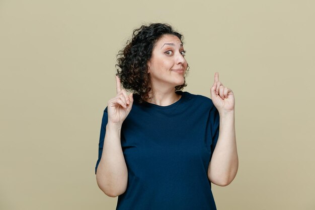 Impressed and pleased middleaged woman wearing tshirt looking at camera pointing fingers up isolated on olive green background