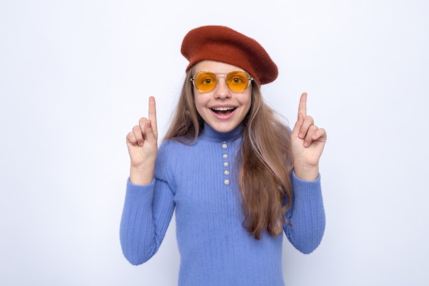 Impressed points at up beautiful little girl wearing glasses with hat isolated on white wall