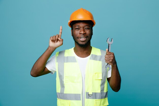 Impressed points at up young african american builder in uniform holding open end wrench isolated on blue background