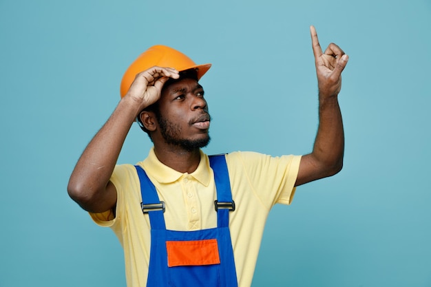 Impressed points at up young african american builder in uniform isolated on blue background