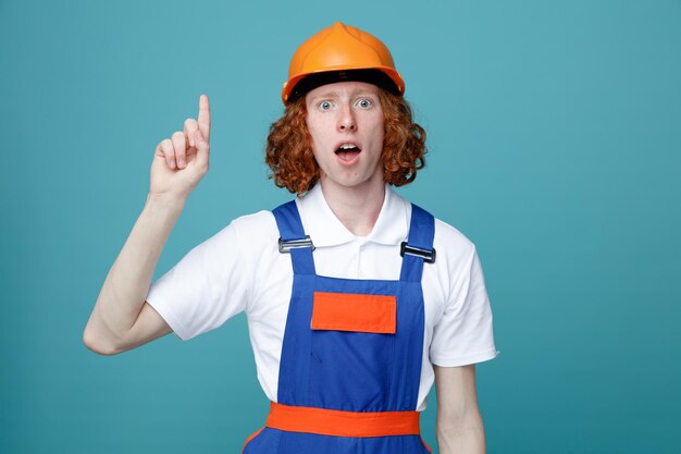 Impressed points at up young builder man in uniform isolated on blue background