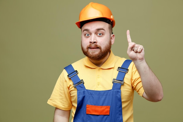 Free photo impressed points at up young builder man in uniform isolated on green background