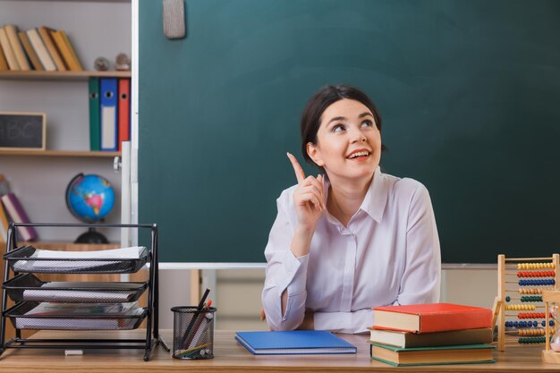 impressed points at up young female teacher sitting at desk with school tools in classroom