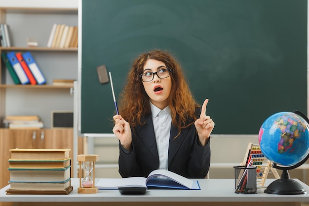 impressed points at up young female teacher wearing glasses holding pointer sitting at desk with school tools in classroom