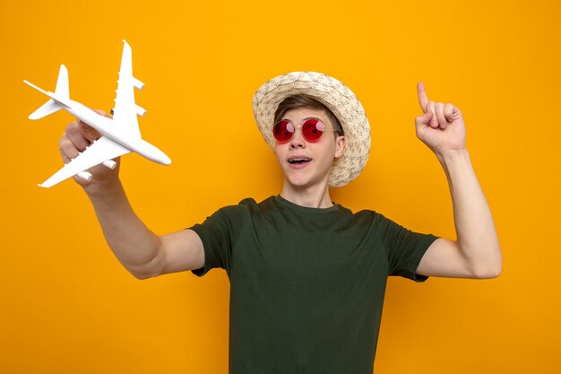 Impressed points at up young handsome guy wearing hat with glasses holding toy airplane 