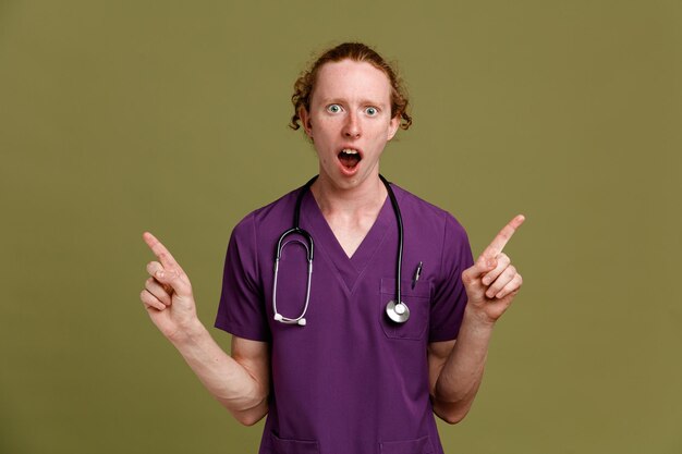 impressed points at up young male doctor wearing uniform with stethoscope isolated on green background