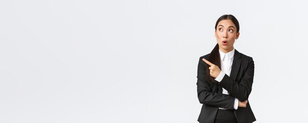 Impressed pretty asian female manager businesswoman in suit pointing and looking upper left corner with amazed expression spot good deal standing white background Copy space