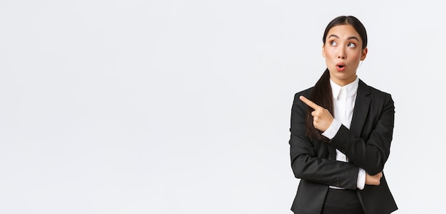 Впечатленная симпатичная азиатская женщина-менеджер, бизнесвумен в костюме, указывая и глядя в левый верхний угол с изумленным выражением лица, замечает хорошую сделку, стоя на белом фоне