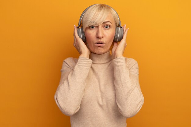 Impressed pretty blonde slavic woman on headphones looks at camera on orange