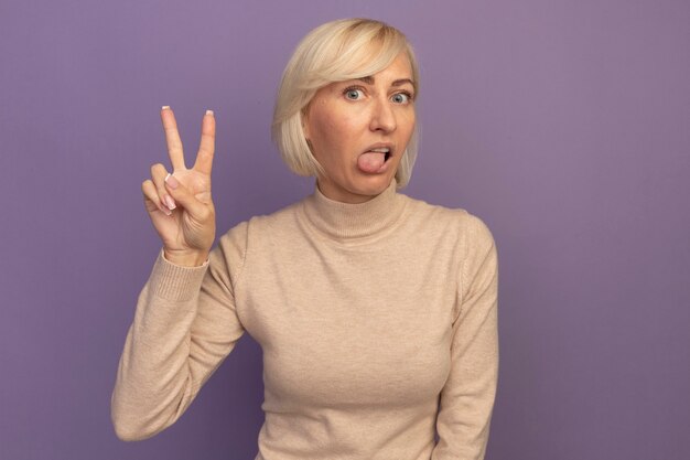 Impressed pretty blonde slavic woman stucks out tongue and gestures victory hand sign on purple
