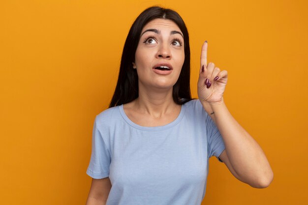 Impressed pretty brunette woman looks and points up isolated on orange wall