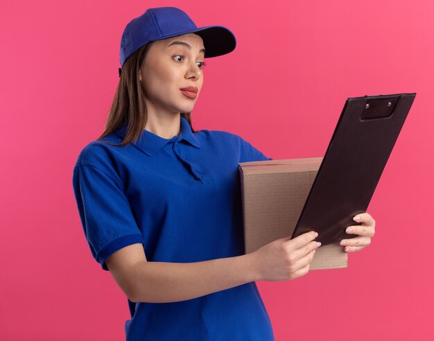 Impressed pretty delivery woman in uniform holds cardbox and looks at clipboard on pink